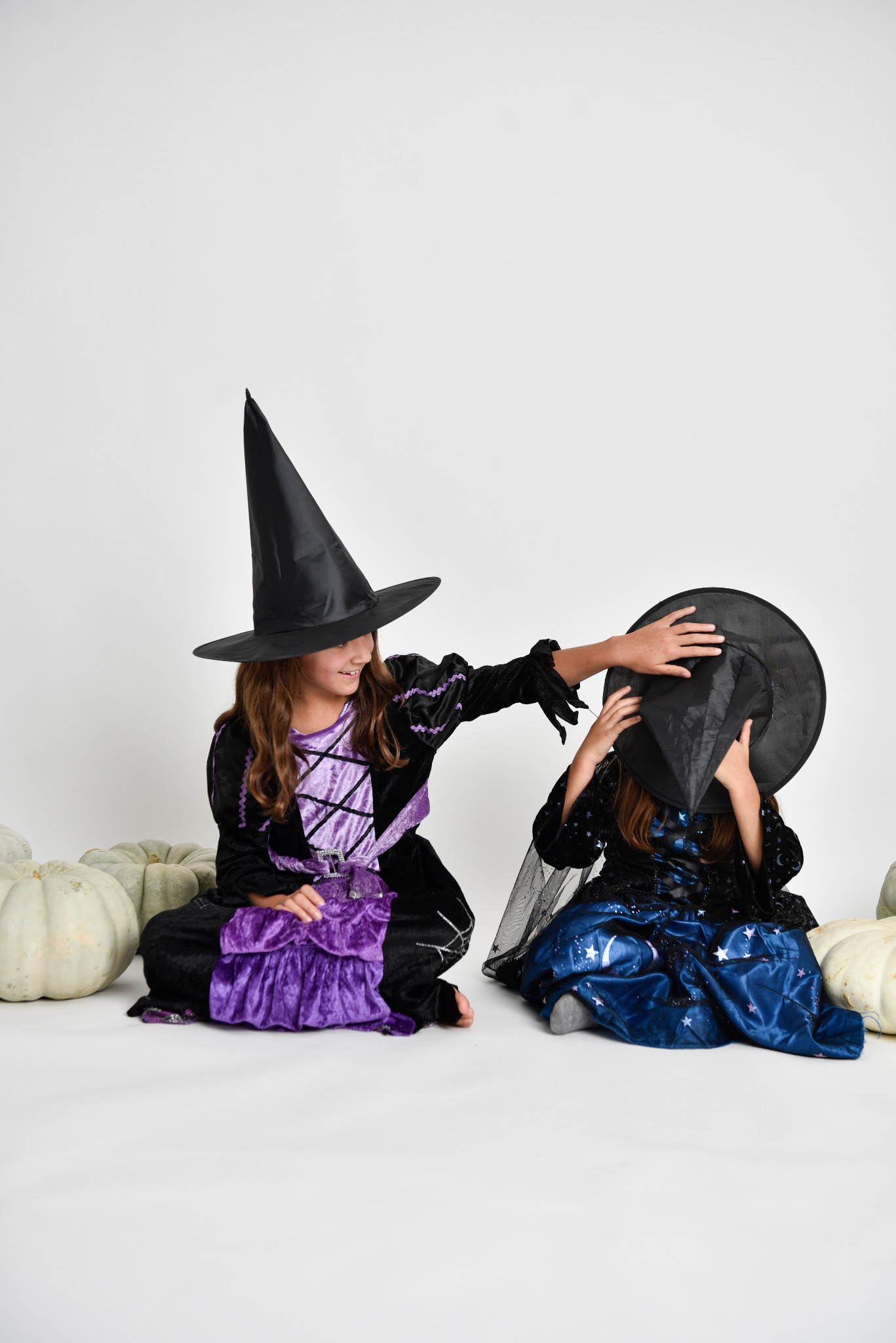 Two children in witch costumes sharing a playful studio moment with white pumpkins
